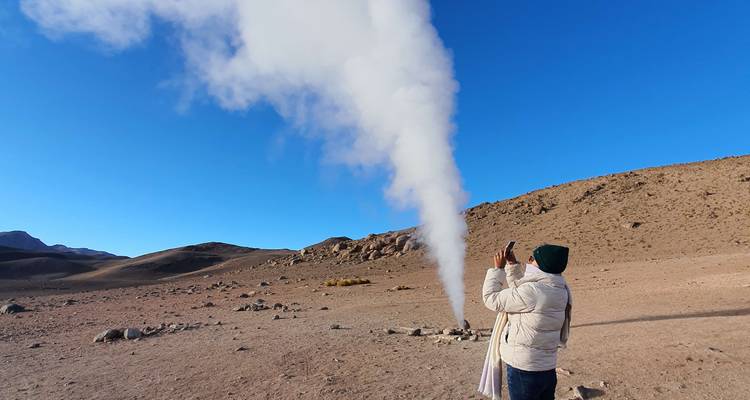 Persoon die een hoge witte stoomwolk fotografeert die opstijgt uit een geothermische bron in een kaal landschap