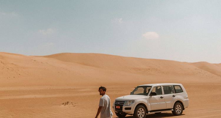 Homme debout à côté d'un 4x4 blanc dans de vastes dunes de sable ondulantes sous un ciel dégagé