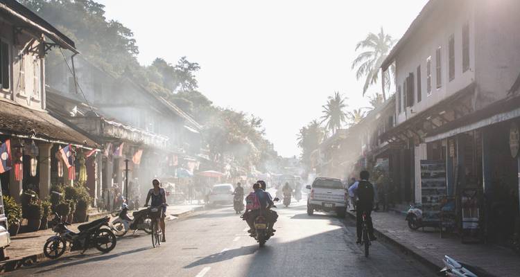 Tegen het licht gefotografeerde ochtendscène op straat met fietsers en scooters die door het mistige zonlicht in Luang Prabang rijden.