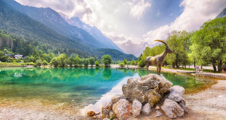 Lac alpin d'un bleu turquoise cristallin, encadré par une forêt et les Alpes Juliennes spectaculaires, avec une statue de bouquetin en bronze sur un rocher au premier plan.