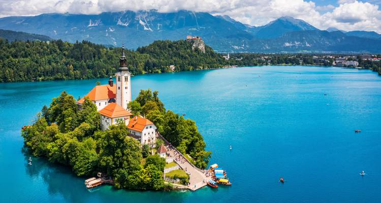 Vue aérienne emblématique du Lac de Bled avec l'église de l'Île de Bled entourée d'une eau bleu vif et des pics alpins au-delà.
