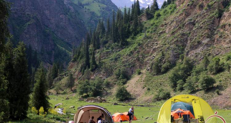 Bergtalcampingplatz mit hell leuchtenden Zelten, die unterhalb steiler, mit Nadelbäumen bewachsener Felswände aufgestellt sind.