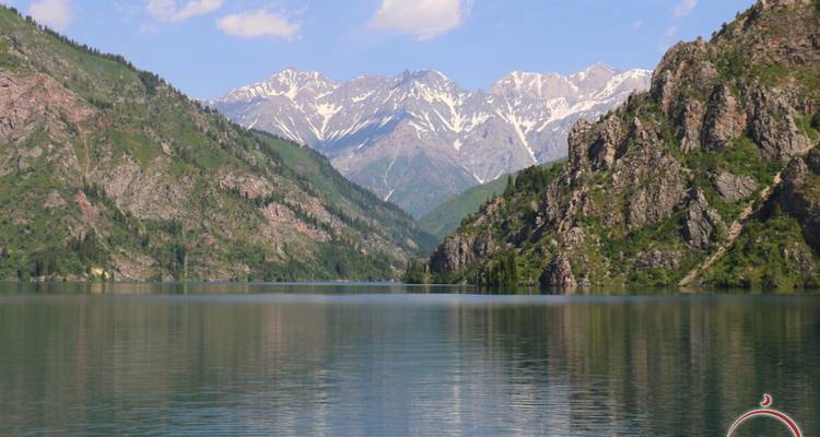 Lago de montaña cristalino que refleja picos nevados y laderas rocosas de color verde.