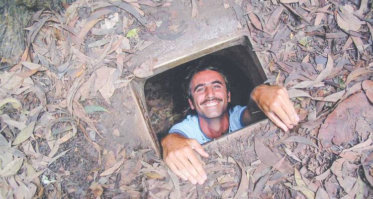 Ein Besucher lächelt, während er aus einem schmalen Cu-Chi-Tunnel auftaucht, der durch trockene Blätter getarnt ist.