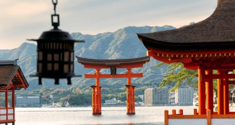 Iconisch vermiljoen torii-poort van het Itsukushima-heiligdom dat uit het water oprijst met bosrijke bergen op de achtergrond.