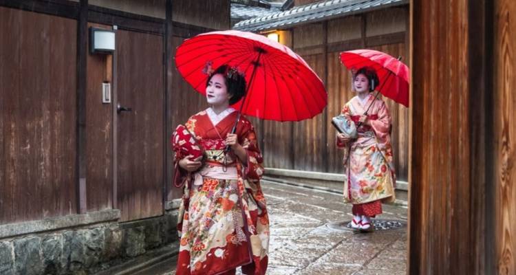 Twee geisha in prachtige kimono's en rode paraplu's lopen door een smal houten steegje op een regenachtige dag in Kyoto.