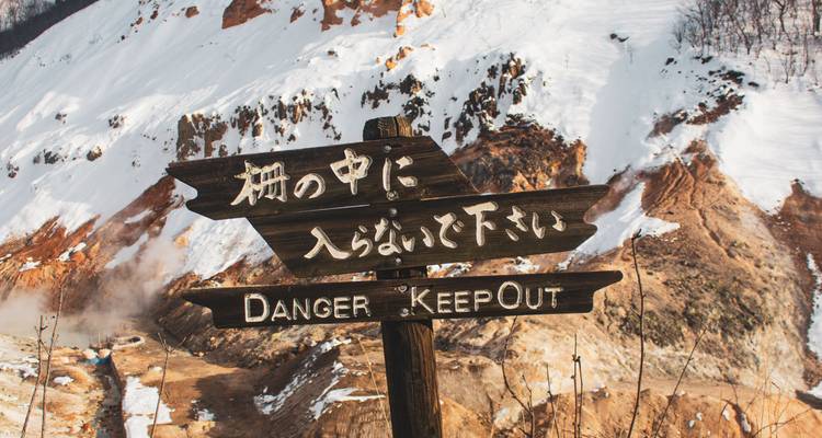 Verwittertes Holzwarnschild in Japanisch und Englisch vor einer schneebedeckten Vulkanlandschaft