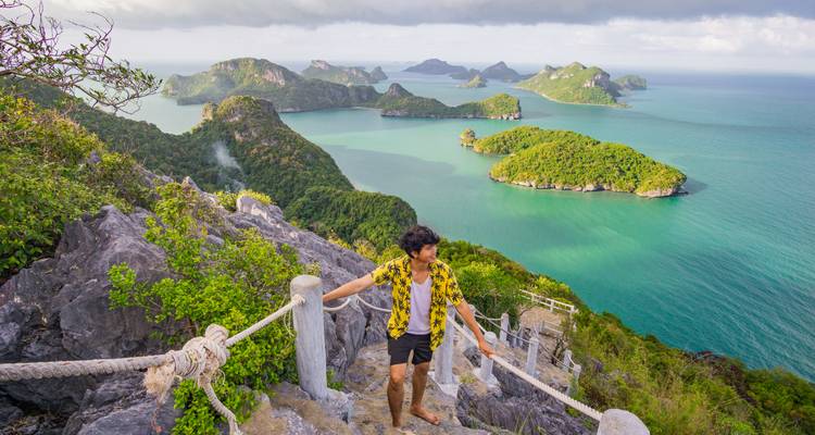 Un voyageur pieds nus monte vers un point de vue rocheux offrant des vues panoramiques sur des îles d'émeraude et une mer turquoise.