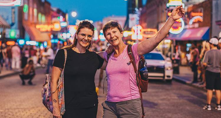 Deux femmes posent joyeusement dans une rue musicale illuminée de néons au crépuscule, entourées de bars animés et d'enseignes lumineuses.