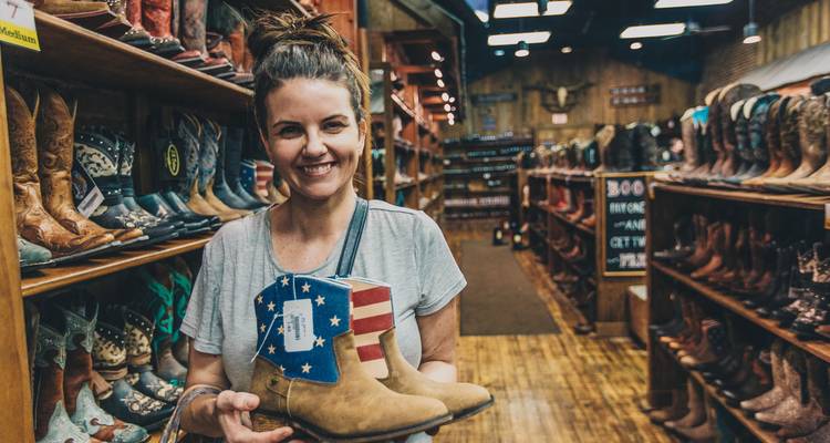 Un client souriant présente une paire de bottes de cowboy patriotiques à l'intérieur d'un magasin de bottes rustique dont les rayonnages sont garnis.