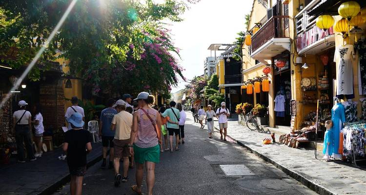 Tourists stroll a lantern-lined street in Hoi An Old Town under bright afternoon sun.