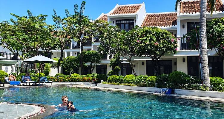 Sunny resort scene with a small family enjoying a swim in a clear blue pool framed by tropical gardens and a white-washed boutique hotel building behind.