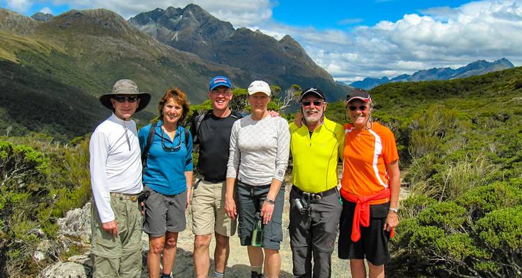 Groupe de randonneurs souriants posant sur un sentier de montagne avec des pics spectaculaires et une vallée verdoyante en arrière-plan.
