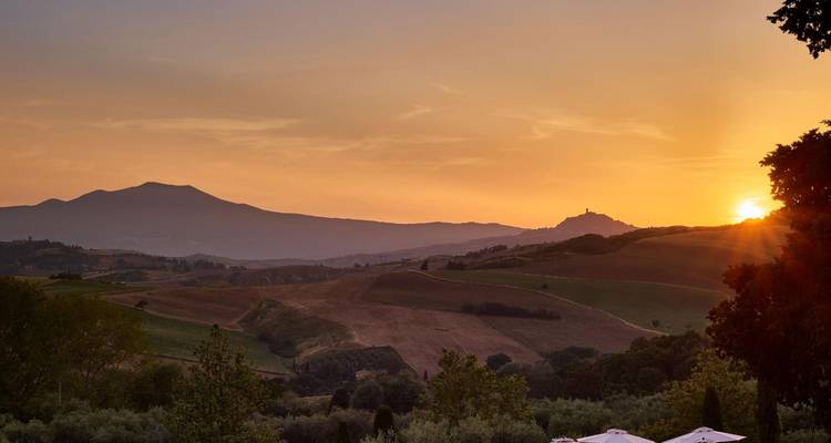 Gouden-uur panorama van rollend Toscaans farmland met een gloeiende zon die achter verre bergkammen verdwijnt.