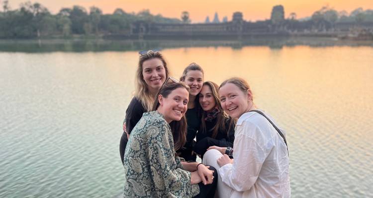 Smiling women seated by a lake at dawn with Angkor Wat’s towers silhouetted in the orange sky.