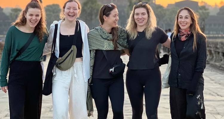 Five friends laughing while walking a stone causeway at sunrise with Angkor’s towers behind.