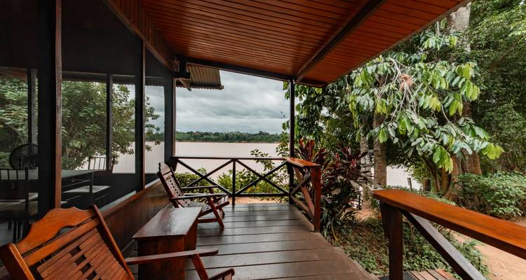 Véranda en bois avec des chaises à bascule et un banc offrant une vue sur un large fleuve de la jungle encadré par une végétation luxuriante.