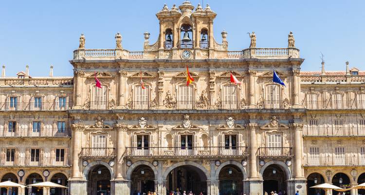 Façade avant de la Plaza Mayor de Salamanque montrant l'élégante tour d'horloge et les drapeaux nationaux sur un ciel dégagé.