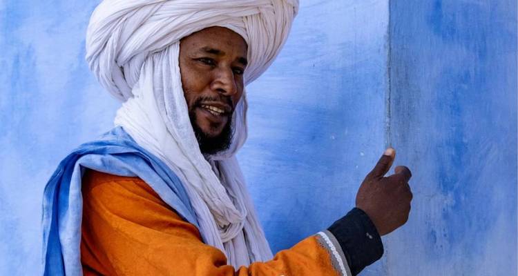 Portrait d'un homme berbère souriant en turban et robe orange devant un mur bleu.