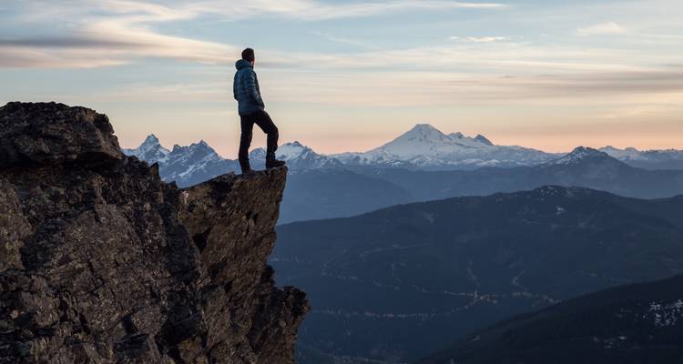 Adventurer standing on cliff edge above expansive mountain landscape at dawn
