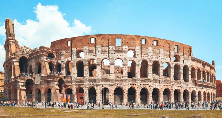 Panoramic daytime view of the iconic Colosseum with crowds of visitors circling its ancient arches under a bright sky.
