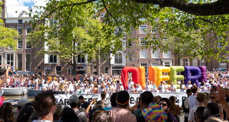 Menschenmassen, die die Amsterdamer Pride-Kanalparade mit bunten QUEER-Buchstaben unter einem belaubten Baum feiern.