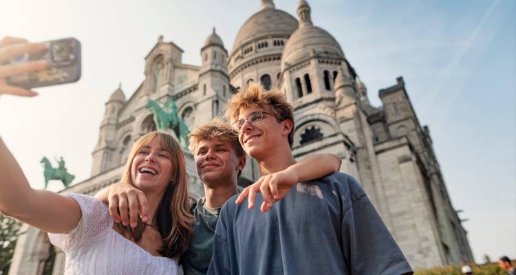 Drei Freunde machen ein Selfie vor den weißen Kuppeln der Basilika Sacré-Cœur an einem hellen Nachmittag.