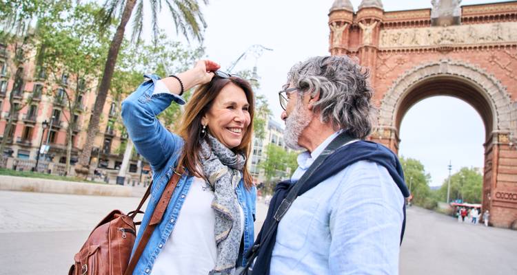 Couple souriant posant près de l'Arc de Triomf de Barcelone avec des palmiers et une arche en brique en arrière-plan.