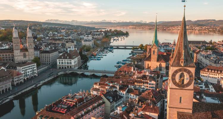 Sunrise aerial of Zurich with church spires and lake backed by snow-capped Alps in the distance.