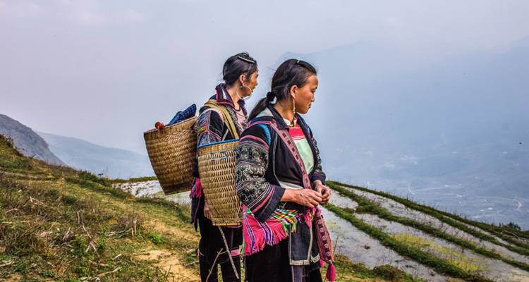 Zwei Hmong-Frauen in traditioneller Kleidung, die über überflutete Reisterrassen und nebelige Täler blicken.