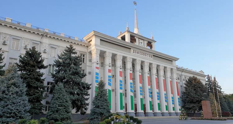 Monumentaal regeringsgebouw in Tiraspol met gekleurde verticale spandoeken en wintergroene bomen ervoor.