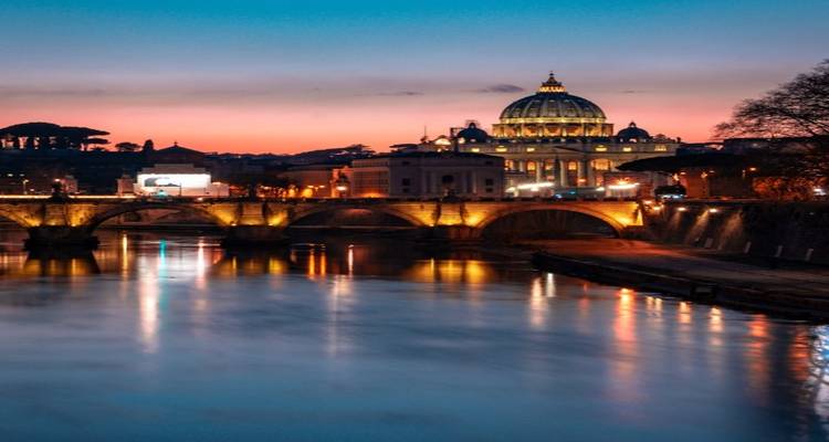 Avondaanzicht van de Sint-Pietersbasiliek en de Tiber die de warme gloed van de stadslichten weerspiegelen.