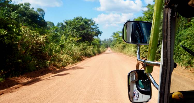 Uitzicht over een lange onverharde landweg vanuit een tuk-tuk, met blauwe lucht en groen gebladerte.