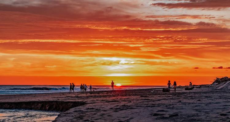 Levendige oranje zonsondergang boven een Stille Oceaan-strand waar mensen wandelen en spelen in de zachte branding.