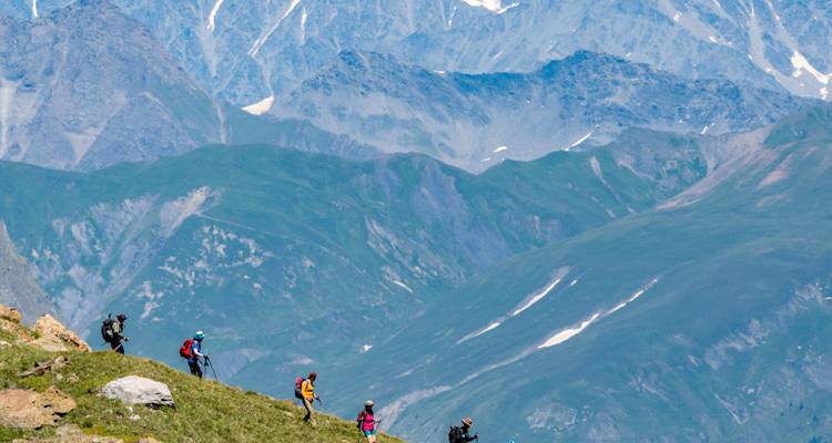 Small group of hikers traverse a grassy alpine ridge with towering grey peaks behind.