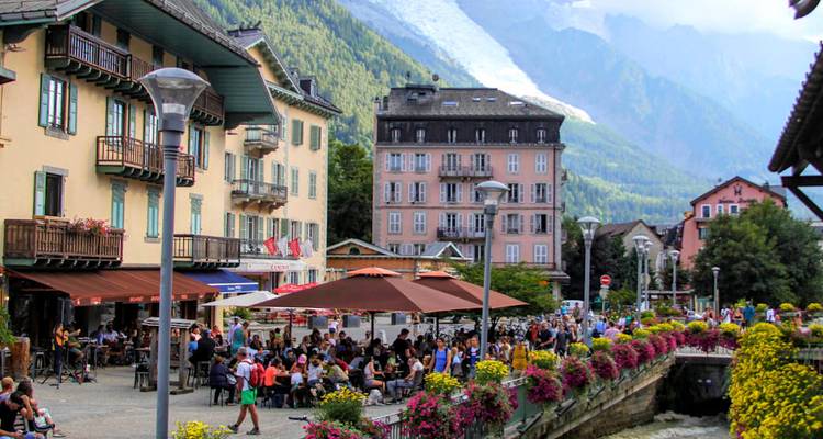 Lively café terraces line a river in Chamonix with snow-capped mountains in the distance.