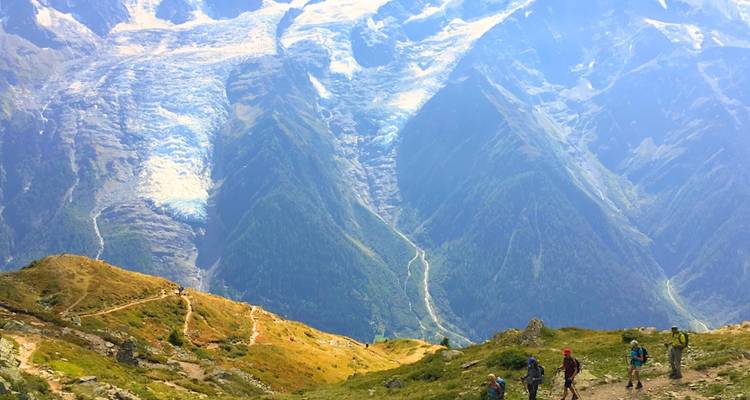 Hikers descend a winding alpine trail with vast glacier-cut valleys stretching below.