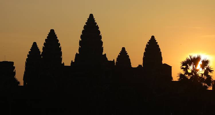 Silhouette of Angkor Wat temple spires against a vivid orange sunrise sky with palm trees.