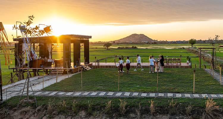Group enjoying sunset at an open-air wooden pavilion overlooking lush green rice fields and distant hill.
