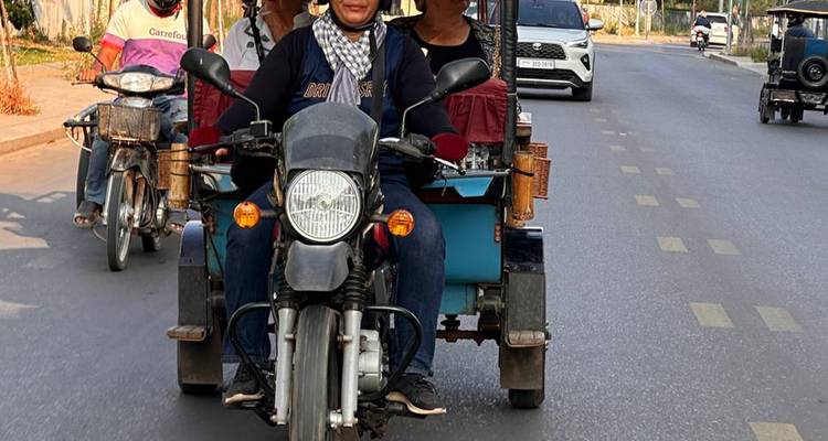 Motorized tuk-tuk with driver and passengers traveling along a paved city road.