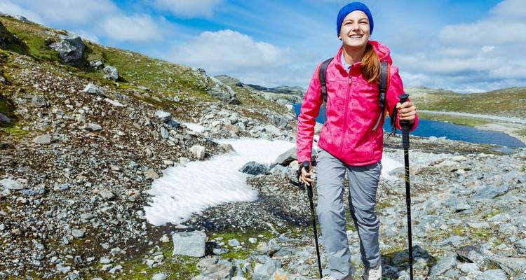 Un randonneur souriant vêtu d'une veste rose vif traverse un terrain alpin rocheux parsemé de plaques de neige.