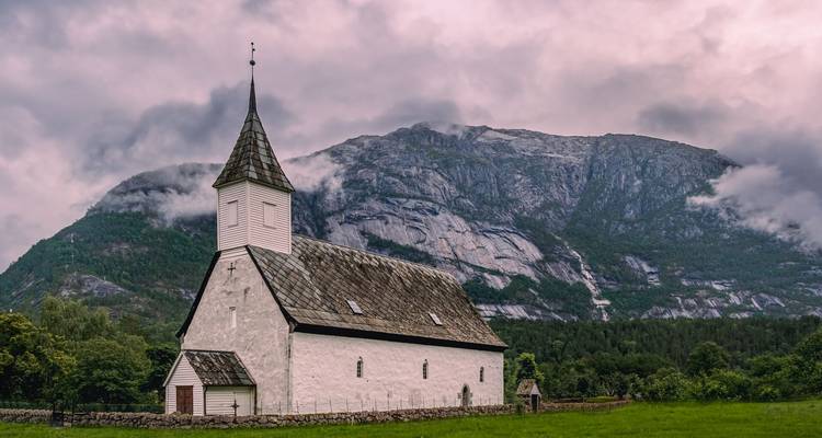 Une église en bois blanc se dresse sur un pré verdoyant sous des montagnes brumeuses avec une cascade.