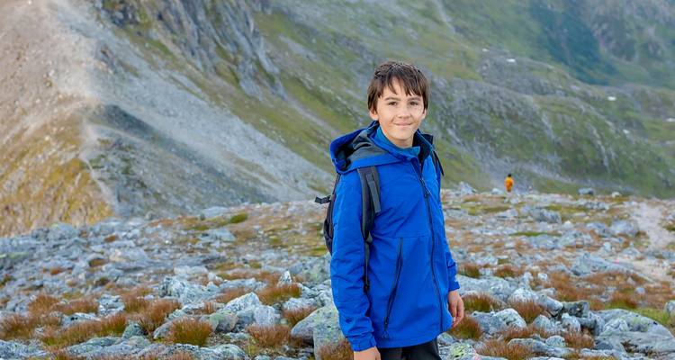 Un garçon vêtu d'une veste bleue se tient sur un versant montagneux rocheux avec des pics escarpés en arrière-plan.