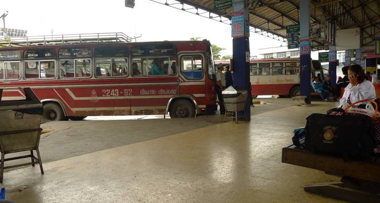 De vieux bus publics rouges garés à l'intérieur d'une gare routière thaïlandaise en plein air avec des voyageurs en attente.