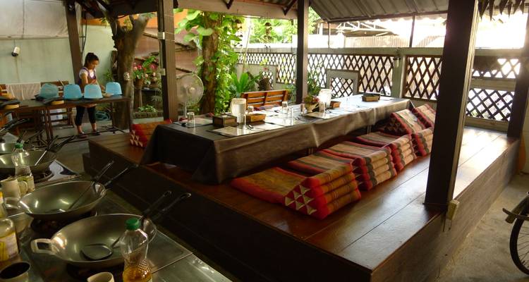 Aménagement traditionnel d'un cours de cuisine thaïlandaise avec des coussins au sol, des woks et un instructeur qui prépare l'espace.