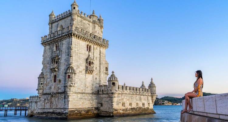 A traveler sits on the riverside wall admiring Lisbon's iconic Belém Tower at dusk.