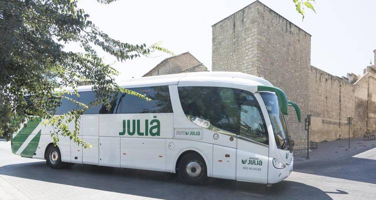 Autobús turístico blanco del grupo Julià estacionado cerca de un muro de piedra antiguo en un día soleado.