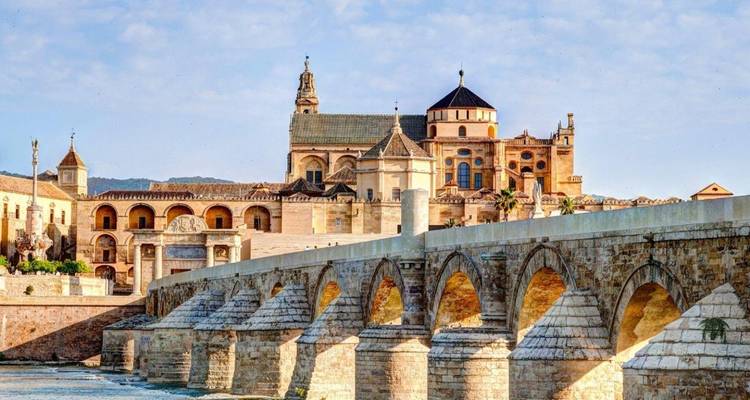 Puente de piedra histórico que conduce al complejo de la Mezquita-Catedral atravesando un río bajo una luz brillante.