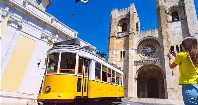 Le célèbre tramway jaune passe devant la Cathédrale de Lisbonne tandis qu'une touriste capture la scène sur son téléphone