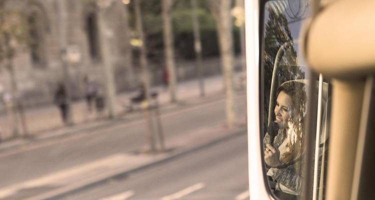 Reflejo de una mujer hablando por micrófono visto en el espejo lateral de un autocar en movimiento.